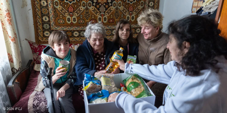 Young man, elderly lady, and two other women sit shoulder to shoulder observing the contents of a food box provided to them by IFCJ Canada