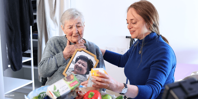 Galina Bailin - daughter was killed by Hamas terrorists on October 7, 2023 eldery woman - light green sweater - holding up photo of her daughter Zinaida - looking at food - hand on her chest Inna Markovitch - adult woman - dark blue jacket - sitting with arm around Galina looking at food box Emergency food box - branded logo - in foreground