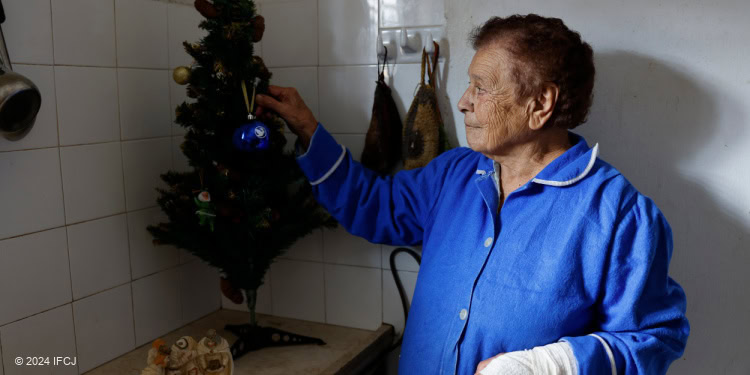 Elderly Leila hangs an ornament on her small Christmas tree on the kitchen counter. Wearing a blue shirt.