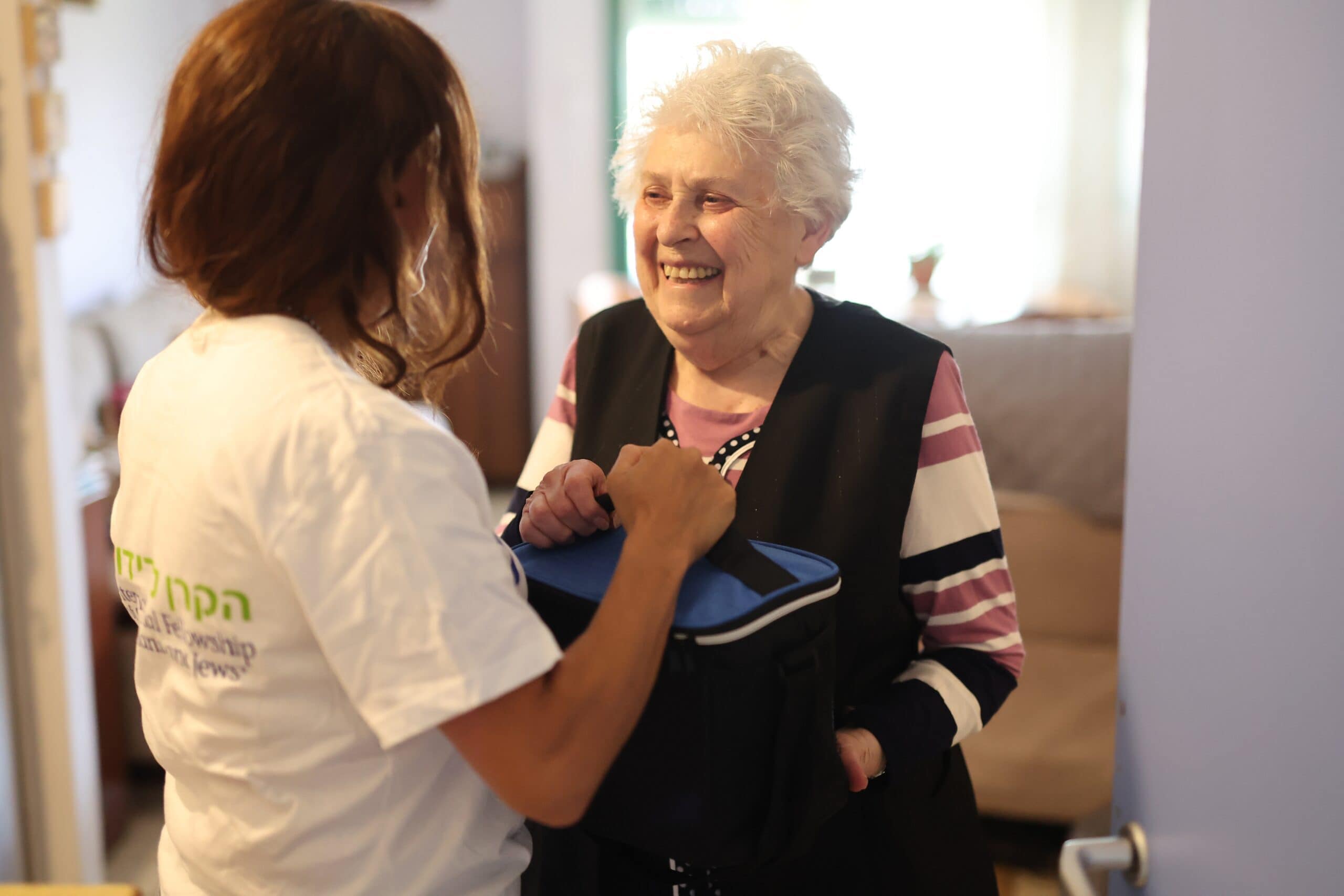 Yevgenya Segal, With Dignity and Fellowship Shavuot 2022, Kiryat Gat, Israel, elderly woman stands with Efrat Klein (IFCJ Staff) holding food bag containing vegetable spreads, fruit jams, and wheat crackers, branded logo tshirt, striped shirt, black pants, black vest, smile