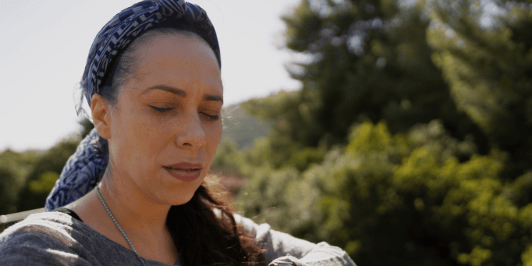 Yael at Nesher Park standing on suspension bridge, close up with eyes closed and leaning on the steel cable of the bridge, trees out of focus in the background, gray sweater