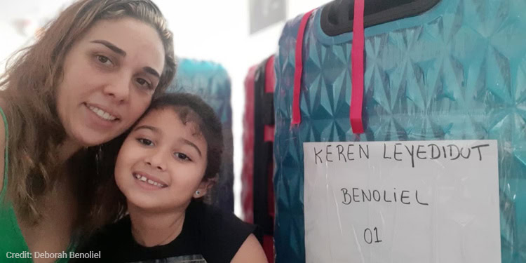Woman and little girl sit next to a suit case with a sign that reads Keren Leyedidut