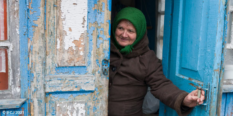 Elderly woman wearing multiple layers of clothing with green scarf on her head, standing in doorway