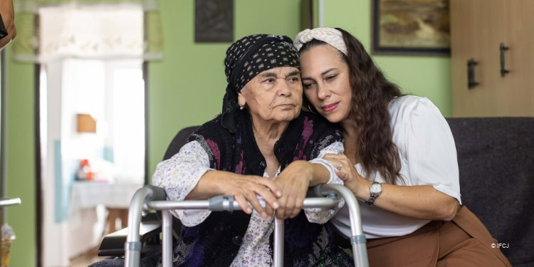 Yael Eckstein and elderly woman wearing a floral nightgown with a walker sit next to each other, head to head.